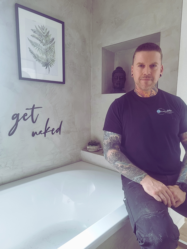 Fugenloses Bad vom Chef persönlich Ein Mann mit Tätowierungen steht neben einer Badewanne in einer modernen Umgebung.